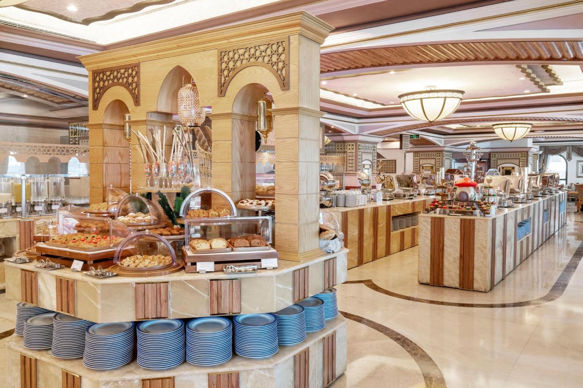 Dar Al Tawhid Intercontinental Makkah By Ihg Hotell Mekka Eksteriør bilde The photo shows a spacious and elegantly designed dining area, likely within a buffet restaurant. In the center, there is a circular display featuring a variety of baked goods and desserts, including pastries and cakes, arranged attractively. Surroun