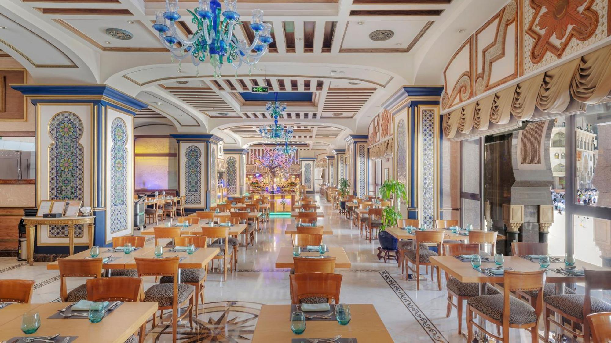 Dar Al Tawhid Intercontinental Makkah By Ihg Hotell Mekka Eksteriør bilde The photo depicts an elegantly designed restaurant interior. It features a spacious layout with numerous wooden tables and chairs arranged neatly. The tables are set with turquoise glassware and tasteful placements. The ceiling is adorned with intric