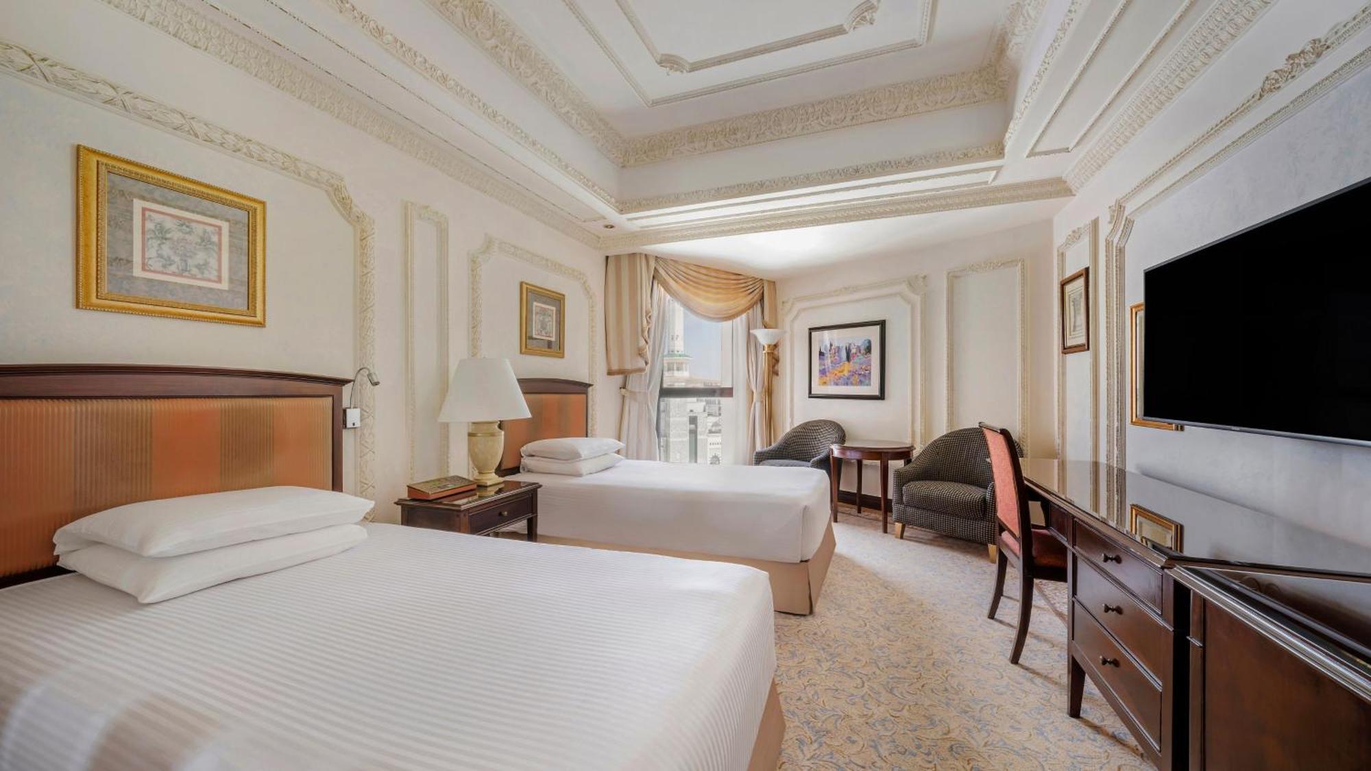 Dar Al Tawhid Intercontinental Makkah By Ihg Hotell Mekka Eksteriør bilde The image shows an elegantly designed hotel room featuring two single beds with white linens. The walls are decorated with light colors and intricate moldings, giving a luxurious feel. There are two bedside tables with lamps beside the beds. A large 